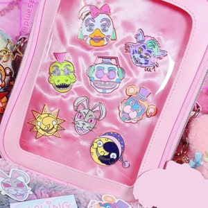 May include: A pink zippered pouch with a clear plastic window displaying a collection of colorful enamel pins featuring cartoon characters. The pins are themed around a popular video game. The pouch is sitting on a fluffy pink and purple surface.