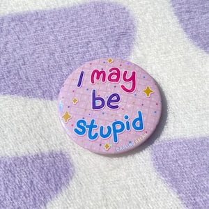 May include: A pink button with a glittery background and white grid pattern. The button says "I may be stupid" in pink, blue, and red letters.