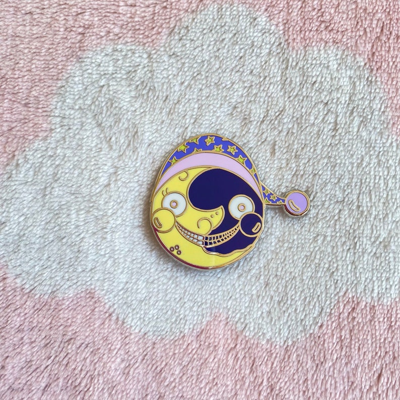 May include: A gold-colored enamel pin featuring a yellow and blue crescent moon with a smiling face. The moon is wearing a purple and blue sleep cap with stars.