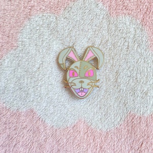 May include: A gold-colored enamel pin featuring a stitched rabbit face with pink ears and pink eyes. The rabbit has a stitched mouth and a pink tongue.
