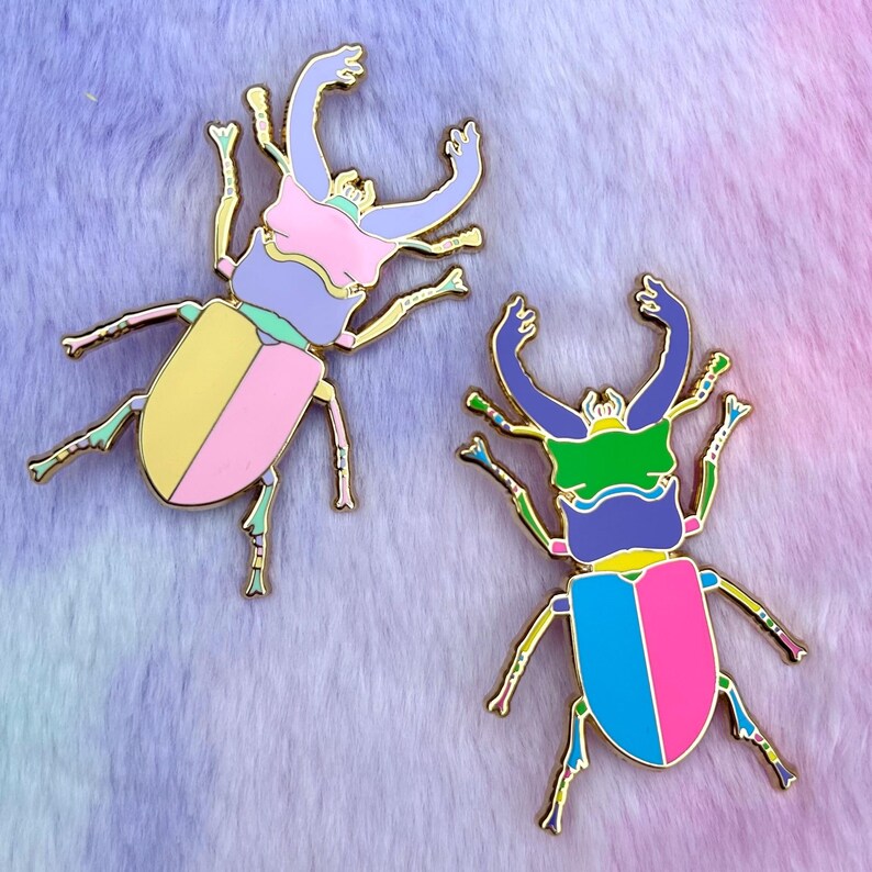 Peut inclure: Deux &eacute;pingles en &eacute;mail color&eacute;es en forme de col&eacute;opt&egrave;res. Le col&eacute;opt&egrave;re de gauche a un corps rose, jaune et violet. Le col&eacute;opt&egrave;re de droite a un corps rose, bleu et vert.