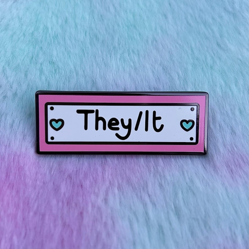 May include: Pink enamel pin with black outline and white background. The pin features the text "They/It" with a blue heart on each side.