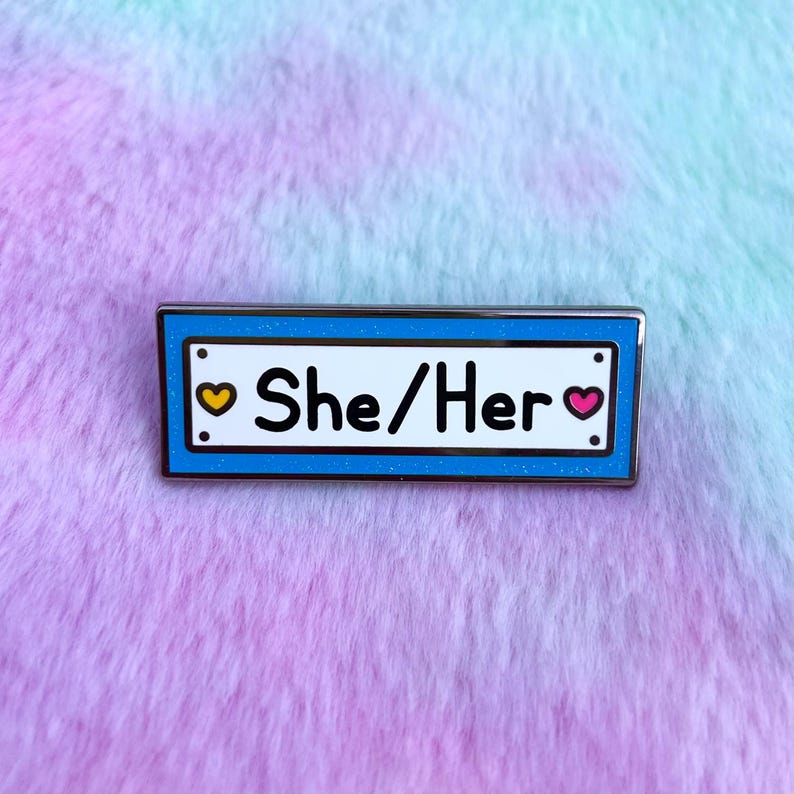 May include: A blue enamel pin with a white border and silver trim. The pin features a white rectangle with the text "She/Her" and a yellow heart on the left and a pink heart on the right.