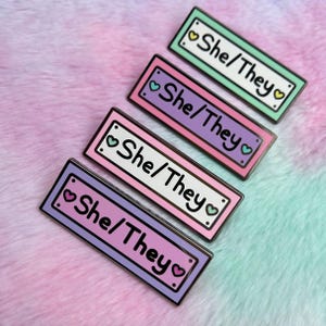 May include: Four enamel pins with a white background and black outlines. Each pin has a different coloured background: green, purple, pink, and light purple. The text on each pin reads "She/They" with a heart on either side of the text.