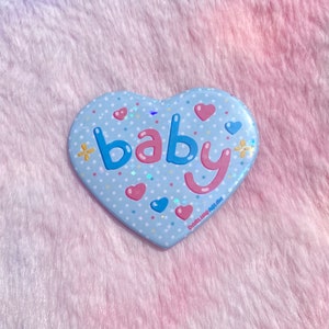 May include: A blue heart-shaped pin with white polka dots and glitter. The word "baby" is written in pink and blue letters with a pink heart and a blue flower on either side.