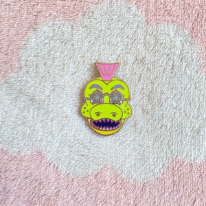 May include: A yellow and green enamel pin of a cartoon dinosaur wearing pink star-shaped sunglasses. The dinosaur has a pink mohawk.