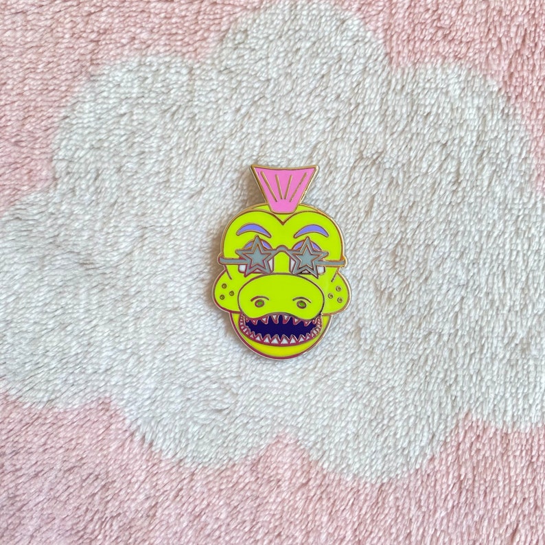 May include: A yellow and green enamel pin of a cartoon dinosaur wearing pink star-shaped sunglasses. The dinosaur has a pink mohawk.