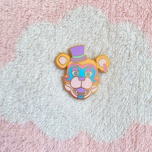 May include: A gold-colored enamel pin featuring a cartoon bear with a purple and gold top hat. The bear has a pink nose, blue eyes, and a purple and blue mouth. The pin is on a pink and white fuzzy surface.