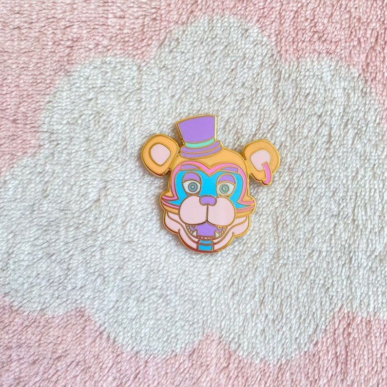May include: A gold-colored enamel pin featuring a cartoon bear with a purple and gold top hat. The bear has a pink nose, blue eyes, and a purple and blue mouth. The pin is on a pink and white fuzzy surface.