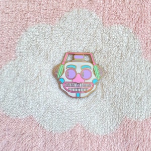 May include: A white enamel pin with a cartoon face wearing headphones. The headphones are green and pink. The face has a pink nose, purple eyes, and a white mouth with teeth.