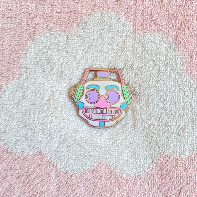 May include: A white enamel pin with a cartoon face wearing headphones. The headphones are green and pink. The face has a pink nose, purple eyes, and a white mouth with teeth.