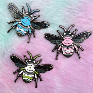 May include: Three enamel pins shaped like bees with glitter accents. Each bee has a different color scheme: blue, pink, and green. The bees have black eyes and a white face with a heart shape.