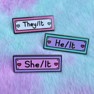 May include: Three enamel pins with a white background and black outlines. Each pin has a different pronoun set: "They/It", "He/It", and "She/It". Each pin has a pink heart on either side of the text.