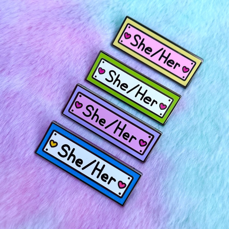 May include: Four enamel pins with a white background and black outline. Each pin has a heart on either side of the text "She/Her" in white. The pins are in different colors: yellow, green, purple, and blue.