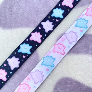 May include: Two fabric ribbon straps with a repeating pattern of cartoon pink, blue, and purple characters. The straps are on a purple and white striped background.
