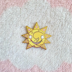 May include: A gold-colored enamel pin featuring a cartoon sun with a crescent moon face. The sun has a pink outline and a yellow face with white eyes and a toothy grin.