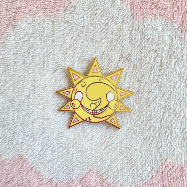 May include: A gold-colored enamel pin featuring a cartoon sun with a crescent moon face. The sun has a pink outline and a yellow face with white eyes and a toothy grin.