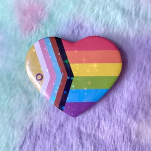 May include: A heart-shaped pin featuring a rainbow flag design. The flag incorporates the colours red, orange, yellow, green, blue, purple, brown, black, and white. The pin has a glittery finish.