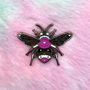 May include: A black and white bee pin with pink accents and glitter. The bee has a heart-shaped body and wings.