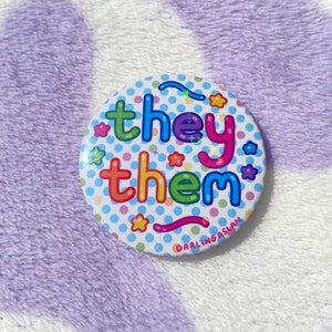 May include: A round button with a white background and colourful polka dots. The words "they them" are written in rainbow colours with a glittery effect. The button has a pink and blue swirl design at the bottom and the text "DARLINGASLAN" in small black letters.