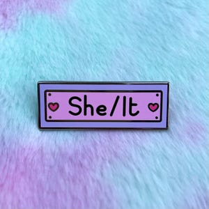 May include: A purple and pink enamel pin with a white border. The pin has two pink hearts on either side of the text "She/It".