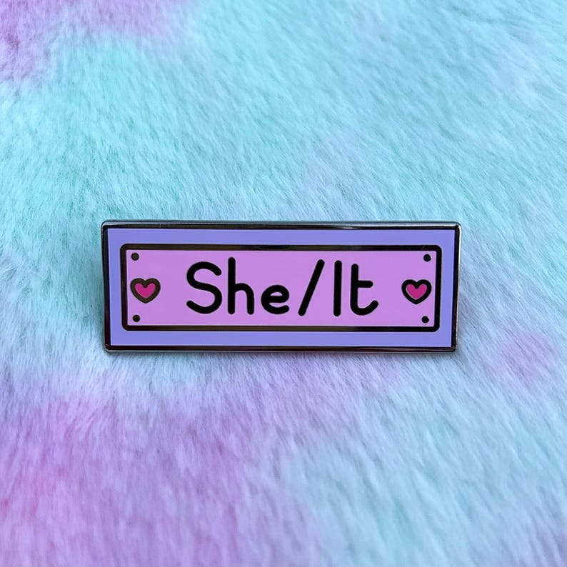 May include: A purple and pink enamel pin with a white border. The pin has two pink hearts on either side of the text "She/It".