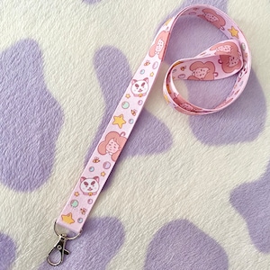 May include: A pink lanyard featuring a white cat and a pink girl character, with stars and bubbles. The lanyard has a silver clip.