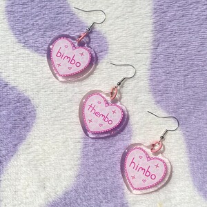 May include: Three pairs of heart-shaped earrings with pink glitter and silver hooks. The earrings have the words "bimbo", "thembo", and "himbo" written on them.