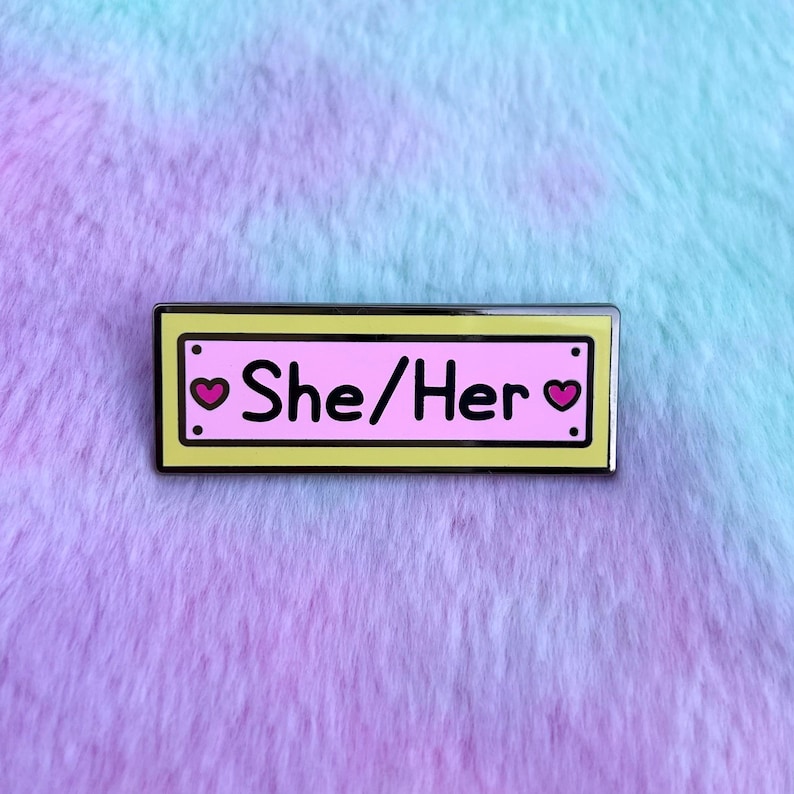 May include: A yellow enamel pin with a pink background and black outline. The pin says "She/Her" with a small pink heart on each side of the text.