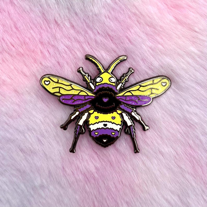 May include: A yellow and purple bee pin with white hearts on the wings. The bee has a black and white striped abdomen and purple and white stripes on the thorax. The bee is on a pink and purple fuzzy background.