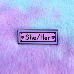 May include: A light purple enamel pin with a silver border. The pin has two pink hearts on either side of the text "She/Her".
