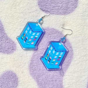 May include: A pair of blue and silver earrings shaped like a TARDIS, the iconic time machine from the Doctor Who television series. The TARDIS is decorated with a variety of pride flags.