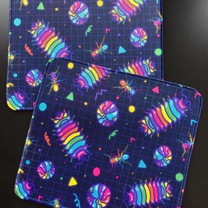 May include: Two square fabric coasters featuring a dark blue grid background with a colourful pattern of rainbow-striped pill bugs, ants, and geometric shapes including circles and triangles in bright neon pink, yellow, green, and light blue.