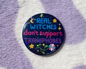 May include: A round, dark blue button with the text "REAL WITCHES don't support TRANSPHOBES" in various colors. The button features stars, a crescent moon, and a flower design. The artist's name, "DARLINGASLAN," is printed at the bottom.