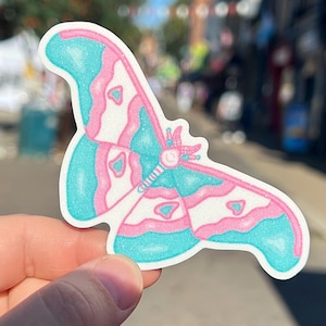 May include: A sticker depicting a stylized butterfly. The butterfly has teal wings with pink and white details. The sticker is held in a hand, with a blurred street scene in the background.