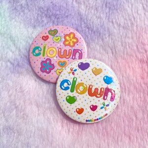 May include: Two round pins with a white background and colourful polka dots. The word "clown" is written in rainbow colours on each pin. One pin has a pink background with a pink flower and hearts. The other pin has a white background with a purple heart, a yellow heart, and a green heart.