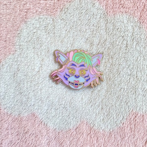 May include: A purple and green enamel pin featuring a cartoon animal with a toothy grin. The animal has pink and blue accents and a green mohawk.