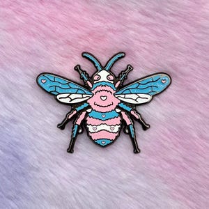 May include: A blue and pink enamel pin of a bee with a trans pride flag pattern. The bee has black legs and antennae.