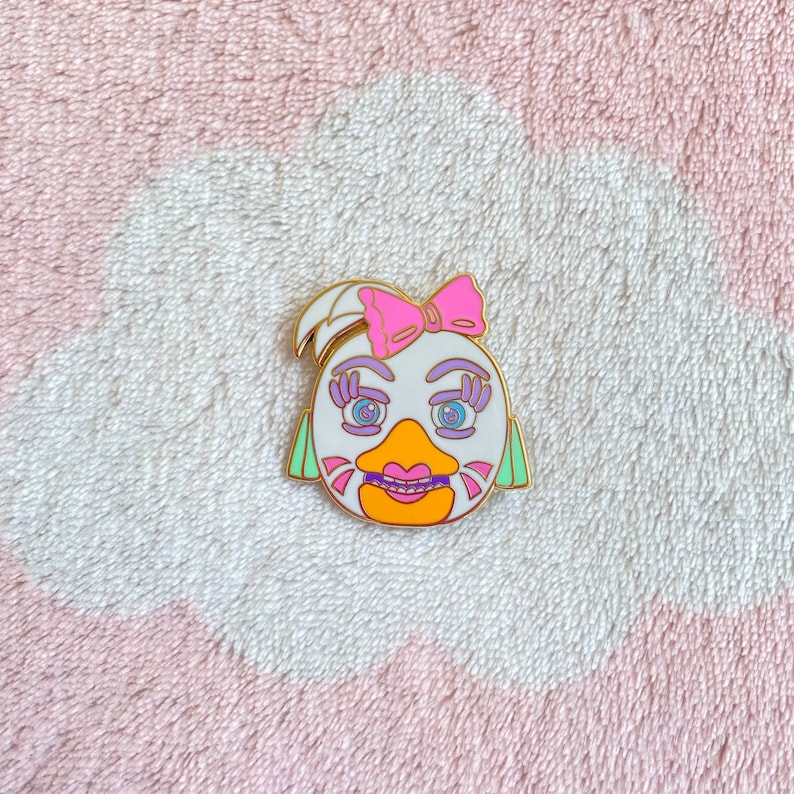 May include: White enamel pin with a cartoon face of a bird with pink eyelashes, purple eyeshadow, blue eyes, a yellow beak, and a pink bow. The bird is wearing braces.