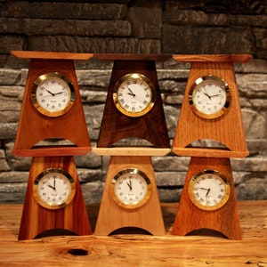 May include: A collection of six wooden table clocks with gold-tone bezels and white faces. The clocks are made of various wood types, including cherry and walnut, and have a unique, angled design. The clocks are displayed on a wooden surface.