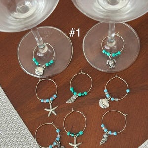 Peut inclure: Charm de verre à vin avec des motifs sur le thème de l'océan. Chaque breloque comporte un anneau argenté orné de perles turquoise et d'un charme, notamment un coquillage, une étoile de mer et une tortue. Les breloques sont exposées sur une surface en bois.