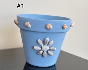 Könnte beinhalten: Hellblau bemalter Blumentopf, verziert mit Muscheln. Der Topf hat ein Muschelblumendesign auf der Vorderseite und kleine Muscheln am Rand. Der Text "#1" ist auf der linken Seite sichtbar.