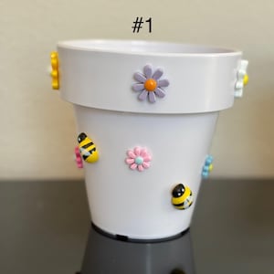 May include: White ceramic plant pot decorated with colourful bee and flower embellishments. The pot has a wide rim and a cylindrical shape. The text "#1" is printed on the pot.