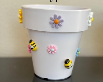 Könnte beinhalten: Weißer Keramik-Blumentopf, verziert mit bunten Bienen- und Blumenverzierungen. Der Topf hat einen breiten Rand und eine zylindrische Form. Der Text "#1" ist auf dem Topf aufgedruckt.