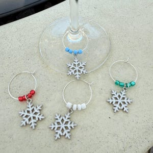 May include: Wine glass charms with snowflake pendants. Each charm features a silver-colored snowflake pendant and a ring adorned with colorful beads. The charms are in red, white, blue, and green.