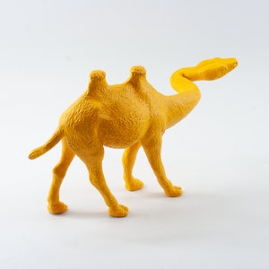 May include: A yellow plastic camel figurine with a long neck and two humps. The camel is standing on a white surface.