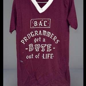 May include: Maroon V-neck t-shirt with white collar and text that reads "B.A.C. Programmers get a -BYTE- out of LIFE" in white lettering. The shirt has a vintage look.