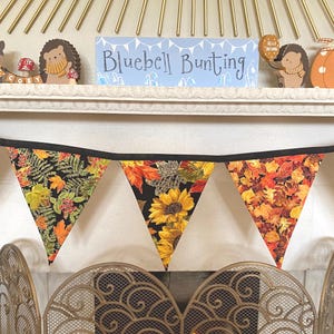 May include: A fabric bunting banner featuring three triangular pennants with autumn-themed patterns. The fabric displays vibrant yellow sunflowers, orange maple leaves, and green foliage on a black background, all attached to a black ribbon.