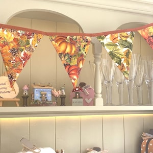 May include: A fabric bunting banner featuring triangular flags with a seasonal autumn print of orange pumpkins, brown pinecones, and multicolored fall leaves, all attached to a solid burnt orange fabric header strip for hanging display.