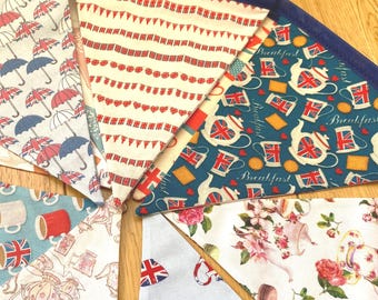 May include: A fabric bunting banner featuring triangular flags with various patterns including red, white, and blue umbrellas, Union Jack flags, teapots, mugs, and floral designs with roses, all connected by a dark blue fabric trim along the top edge.